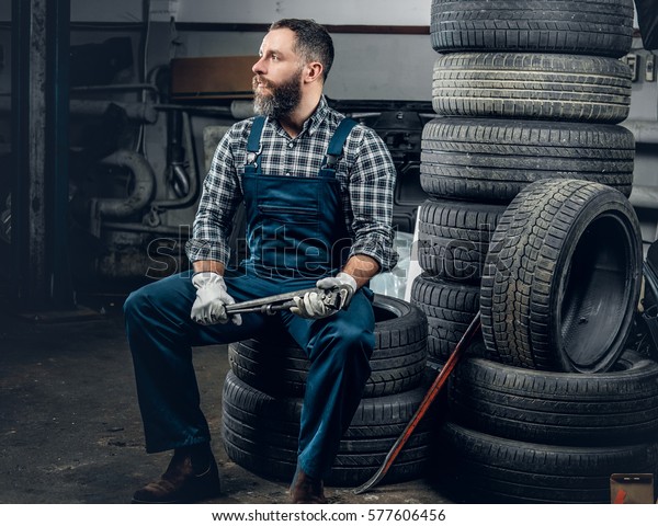 The brutal bearded mechanic male sits on an old\
tire in a garage.