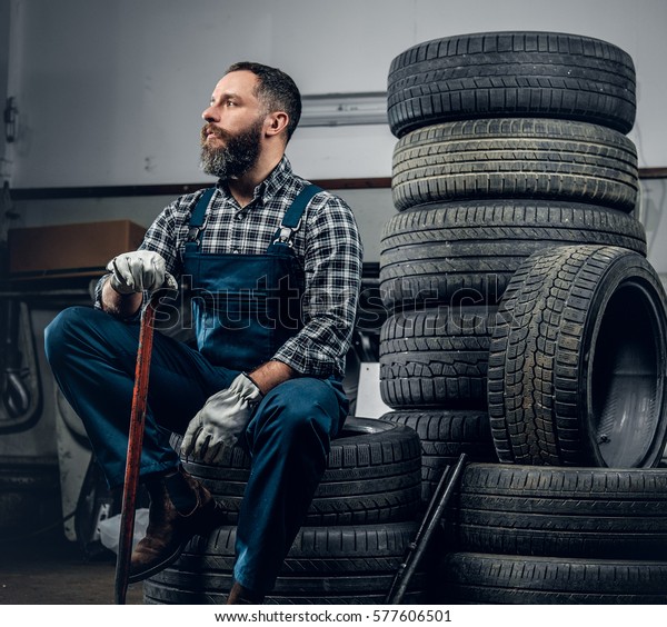 The brutal bearded mechanic male sits on an old\
tire in a garage.