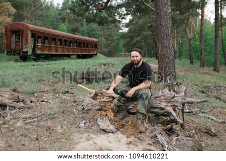 Foto Bild bärtiger Holzfäller mit einer Axt, die neben einem rostigen Zug sitzt.