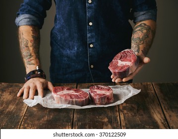 Brutal Butcher With Tattooed Hands Offers A Piece Of Great Raw Meat Steak On Camera, Other Steaks In White Craft Paper On Vintage Old Wooden Grunge Table. Unrecognizable