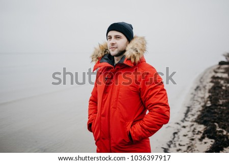 A brutal man with a beard in the red parka, black cap, and blue jeans stands on the beach and looks at the sea. A sailor in a parka on the shore waiting for a ship. Jurmala Latvia.
