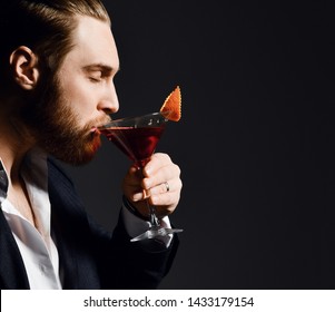 Brutal modern man in fashion suit bearded barman with closed in pleasure eyes drinks a delicious red cocktail with orange in martini glass on dark background. - Powered by Shutterstock