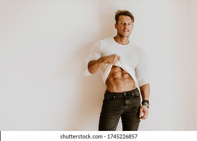 Brutal muscular man showing six pack abs muscles indoors. Brutal muscular man wearing white shirt showing abs muscles. Muscles and abs. Joyful male showing abs stomach at home wearing casual jeans