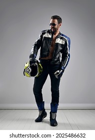 Brutal, Self-confident Adult Man Biker Motocross Racer In Motorcycle Leather Gear High Boots, Jacket And Gloves Walks With His Helmet In Hand, Looking Aside Over Light Background. Full-growth