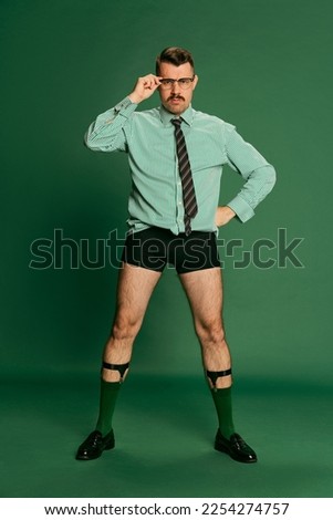 Brutal, serious boss. Portrait of handsome man, businessman standing in shirt without pants, posing over green studio background. Concept of emotions, business, occupation, facial expression, fashion