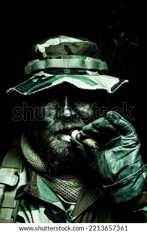 Brutal and serious commando soldier, army special forces veteran, camouflage battle uniform, boonie hat, black paint on bearded face, combat knife in shoulder holder, smoking cigar, studio portrait