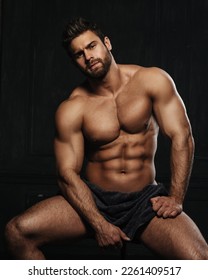 Brutal sexy man with six pack abs in towel at black background. Handsome fitness male model sitting in studio. Bearded muscled guy covering with towel in studio.