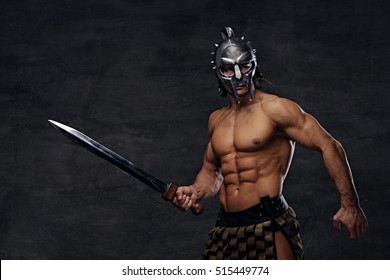 Brutal shirtless muscular male in a silver gladiator helmet holds an iron sword on grey background in a studio.