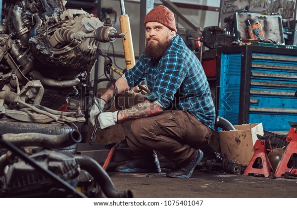 A brutal tattooed bearded mechanic specialist\
repairs the car engine which is raised on the hydraulic lift in the\
garage. Service station.