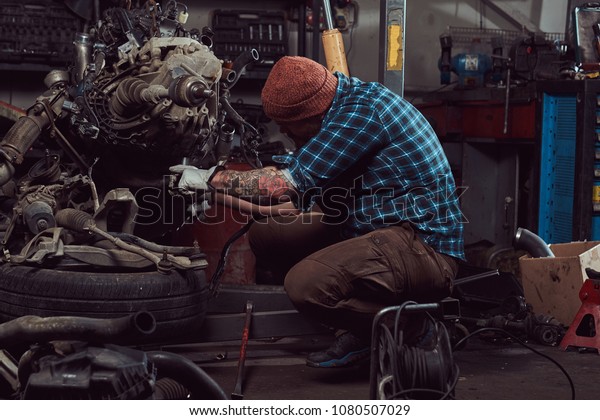 A brutal tattooed bearded mechanic specialist\
repairs the car engine which is raised on the hydraulic lift in the\
garage. Service station.
