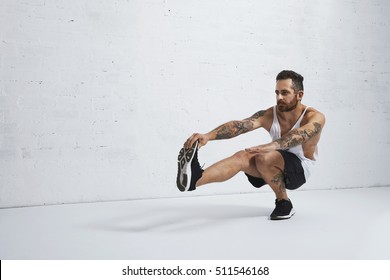 Brutal tattooed calisthenics coach shows exercise moves one leg squat, isolated on white brick wall