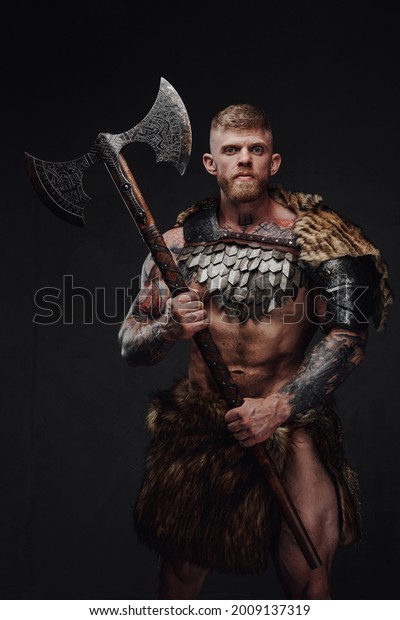 Brutal tattooed warrior wearing light
armour and fur holding two-handed axe in dark
studio