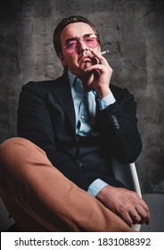 Brutal Unshaved Haughty Man With Slicked Back Hair In Stylish Official Suit And Smoked Blue Glasses Sits With His Legs Crossed Smoking Cigarette Over Dark Background. Stylish Look For Men Concept