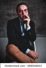 Brutal Unshaved Man With Slicked Back Hair In Stylish Official Suit And Smoked Blue Glasses Sits With His Legs Crossed Smoking Cigarette Over Dark Background. Stylish Look For Men Concept