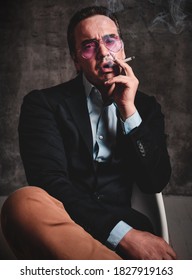 Brutal Unshaved Manwith Slicked Back Hair In Stylish Official Suit And Smoked Blue Glasses Sits Smoking Cigarette, Blowing Out Over Dark Background. Stylish Look For Men Concept