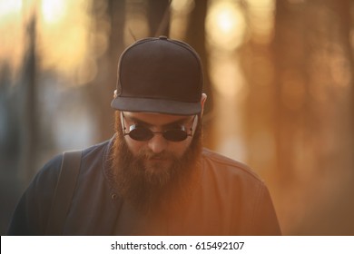 A Brutal Young Man With A Huge Beard In Sunglasses And A Cap In The Woods At Sunset. Lifestyle.