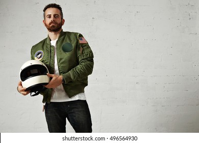 Brutal Young Man In Military Jacket With Embroidery Patches Holds His Motocycle Helmet In Hands And Proudly Looking On Camera, Isolated On White Wall With Bricks