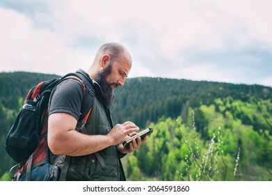 A Brutal Young Man Stands By The Mountains And Looks At A Mobile Phone. The Hiker Is Looking For A Road On The Map. Outdoor Beauty. Freedom Concept. Enjoy, Copy Space.Side View.