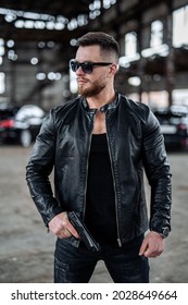 Brutal Young Man In Total Black Outfit And Sunglasses Points A Gun On Some Point. Abandoned Blurred Background Of Warehouse