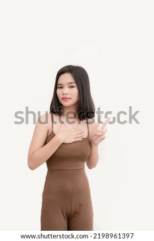 A bubbly woman wearing a brown tank top with one hand on chest and the other showing peace sign isolated on a white background