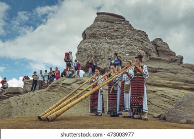 Bucegi Mountains, Romania - August 6, 2016: Young Romanian female artists wearing colorful traditional costumes play the tulnic near the legendary Sphinx megalith in Bucegi mountains..