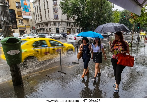 Bucharest, Romania - August 01, 2019: Women walking\
with umbrellas, in rainy weather, while a taxi splashes them with\
water from a puddle in downtown Bucharest. This image is for\
editorial use onl