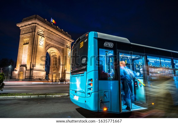 Bucharest, Romania-February 14th 2020:The\
arch of Triumph, Triumph Arch. Night traffic in front of the\
Triumphal Arch. People getting on the bus. Bucharest transport\
service. Long exposure\
photography