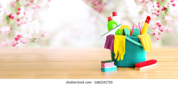 Bucket or basket with cleaning items on wooden table with spring flowers background. Washing set with copy space banner.