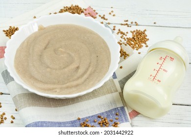 Buckwheat Porridge For A Baby From Ground Cereals In A White Bowl, A Bottle Of Milk On A White Wooden Background, Side View. The First Complementary Food Of A Child, Baby Nutrition.