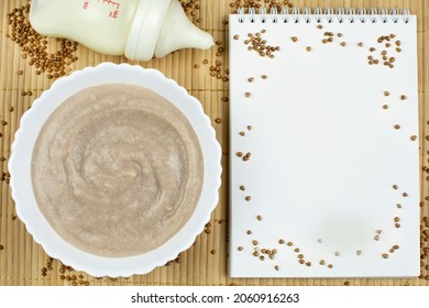 Buckwheat Porridge For The Baby From Ground Cereals In A White Bowl, A Bottle Of Milk, A Notebook With A Blank Page For Writing On A Brown Background. Baby's First Complementary Food, Baby Nutrition. 