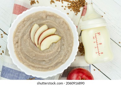 Buckwheat Porridge For A Baby Made Of Ground Cereals In A White Bowl With An Apple, A Bottle Of Milk On A White Wooden Background. Baby's First Complementary Food, Baby Nutrition.