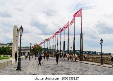 Budapest, Hungary, 6 August 2019: Many Tourists Walking Near Vivid Colorful Flags Blowing In The Wind In Direct Sunlight Towards Cloudy Blue Sky In A Sunny Day Near The Buda Castle District