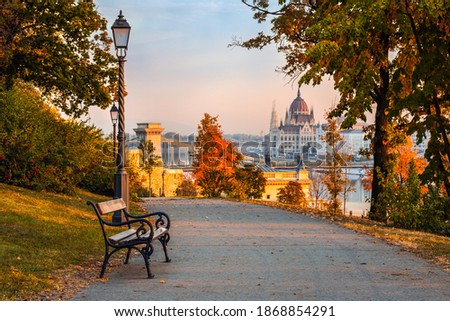 Budapest, Hungary - Bench at Varkert Bazaar with lamp post, autumn foliage, Szechenyi Chain Bridge and Parliament at background on a warm autumn morning