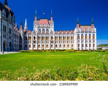 Budapest, Hungary - July 17th 2022:Hungarian Parliament Building With Hungarian Flag On It. East Façade Of The Building With Green Lawn With Flowers In First Plan And Clear Blue Sky In The Background.