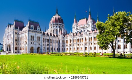 Budapest, Hungary - July 17th 2022:Hungarian Parliament Building With Hungarian Flag On It. East Façade Of The Building With Green Lawn With Flowers In First Plan And Clear Blue Sky In The Background.