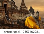 Buddha with golden robe against blurred Central Chedi of Wat Yai Chai Mongkol background in Ayutthaya, Thailand. Buddhist postcard with copy space. Buddha with yellow scarf view from the back