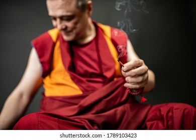 Buddhist monk in red kesa, holding an incense stick with incense in his hand