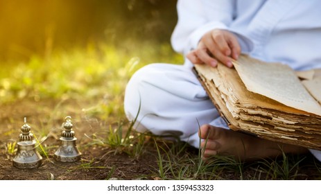 Buddhist  monk sits in  yoga lotus position meditating, reading old open book of wisdom, and there are a number  Tibetan bells and burn incense at meadow. beautiful sunset background