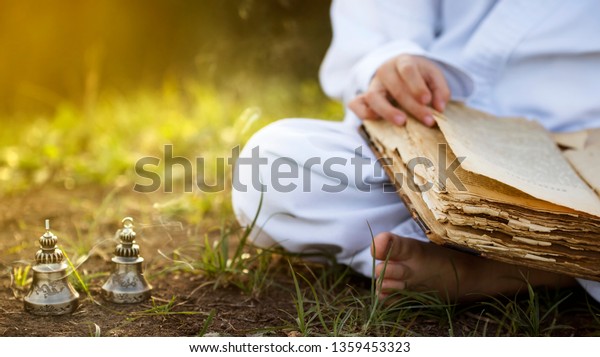 Buddhist \
monk sits in  yoga lotus position meditating, reading old open book\
of wisdom, and there are a number  Tibetan bells and burn incense\
at meadow. beautiful sunset\
background