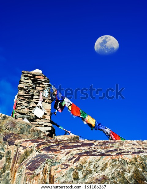 Buddhist stone tower with praying flags\
and moon at Himalaya high mountain road pass at Manali - Leh\
highway over blue sky. India, Ladakh, altitude\
4500m
