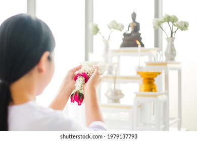 Buddhist woman wearing Thai traditional white dress and holding jasmine garland for praying to buddha statue on Holy day. Asian Female worship and meditation at home .