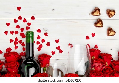 Buds of red roses and bottle of wine on white wooden background with confetti, glass and chocolate