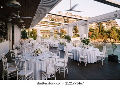 Budva, Montenegro - 14.07.21: Wedding dinner table reception. Lots of round tables with white tablecloths and white Chiavari chairs. Restaurant without roof overlooking modern villa complex 