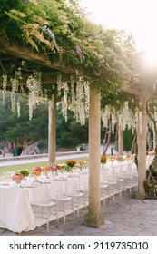 Budva, Montenegro - 15.07.21: Wedding dinner table reception. A very long table for guests with a white tablecloth, chairs Chiavari. Under the old columns with vines of wisteria.