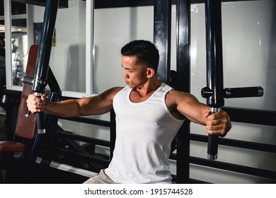 A Buff Asian Guy In A White Tank Top Uses The Seated Lever Fly Machine At The Gym. Working Out Chest Muscles.