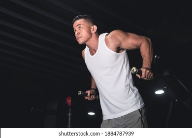 A buff asian man in a white tank top begins to do some tricep dips on the dip station. Working out triceps and arms at the gym.