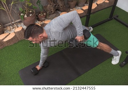 A buff man does a set of renegade rows with hex dumbbells. Working out back muscles and core at his home gym.