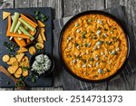 buffalo chicken dip in baking dish on grey rustic wooden table with crackers, blue cheese, carrot and celery sticks on slate board, horizontal view from above, flat lay