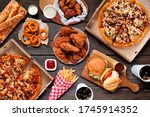 Buffet table scene of take out or delivery foods. Pizza, hamburgers, fried chicken and sides. Above view on a dark wood background.