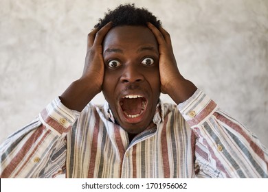 Bug eyed shocked young Afro American man in panic holding hands on head and yelling, stressed with alarming news, being worried about world situation. Black guy having terrified look, screaming
